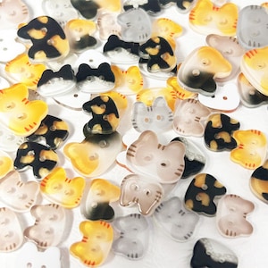 May include: A collection of cat-shaped buttons in various colors and patterns, including orange, black, white, and gray. Each button features two holes for sewing and depicts a stylized feline head with small ears and subtle whisker markings.