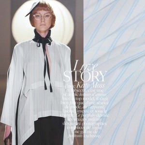 May include: A woman wearing a white shirt with a black tie and a black skirt. She is wearing a hat with a brim and glasses. The background is a light blue and white striped fabric. The text "Love Story" is written in white letters on the fabric. The text "Entre Kate Moss et l'objectif, c'est une veritable histoire d'amour. Avec un top model, il s'agit bien plus que d'une s&eacute;ance de mode, dit l'appareil, fascine, inspire et sous le charme. Ce portfolio d&eacute;voile les images pr&eacute;f&eacute;r&eacute;es de quinze photographes complices de Vogue. Une galerie de portraits exclusive." is written in black letters on the fabric.