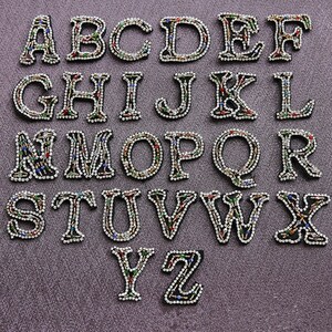 Pode incluir: Um conjunto de 26 letras do alfabeto para passar a ferro, cada uma decorada com strass coloridos. As letras são maiúsculas e estão dispostas em ordem alfabética.