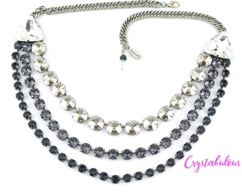 May include: A multi-layered necklace with a silver chain and three rows of crystal stones. The top row features larger clear crystals, while the bottom two rows feature smaller, darker gray crystals.