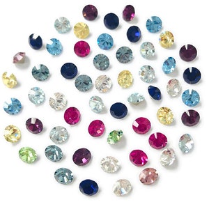 May include: Assortment of colorful rhinestones in various shapes and sizes, including round, square, and oval. The rhinestones are arranged in a circular pattern on a white background.