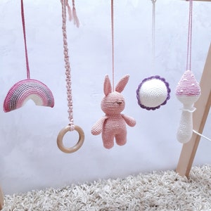 May include: A wooden baby play gym with five hanging toys: a pink and purple rainbow, a pink crocheted bunny, a pink and purple crocheted flower, a pink crocheted ring, and a pink and purple crocheted rattle.
