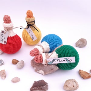 Puede incluir: Cuatro botellas de poci&oacute;n de crochet en rojo, amarillo, azul y verde con detalles blancos. Cada botella tiene una peque&ntilde;a etiqueta con la palabra "Poci&oacute;n" y un n&uacute;mero.