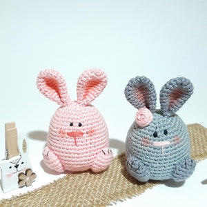 May include: Two crocheted bunny plushies, one pink and one gray, with embroidered faces. The plushies are sitting on a burlap surface with wooden bunny clothespins on either side.