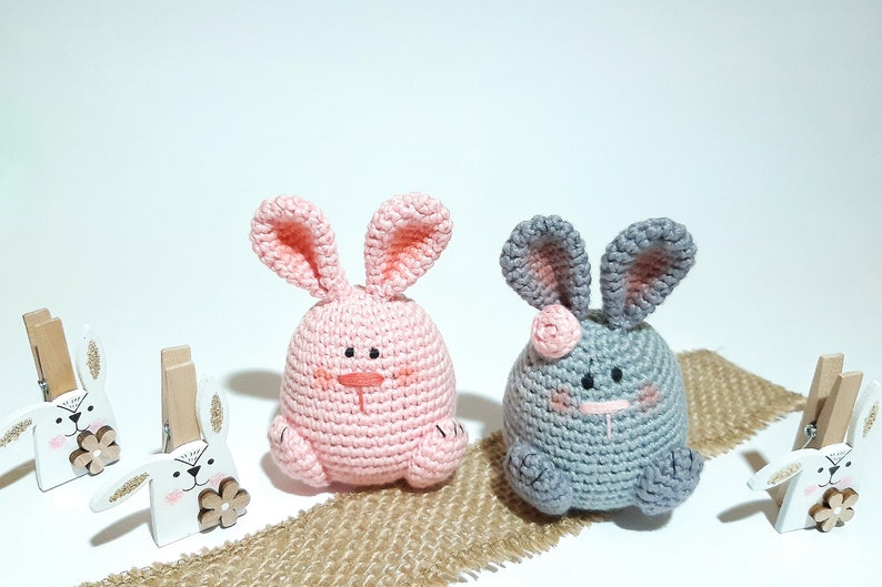 May include: Two crocheted bunny plushies, one pink and one gray, with embroidered faces. The plushies are sitting on a burlap surface with wooden bunny clothespins on either side.