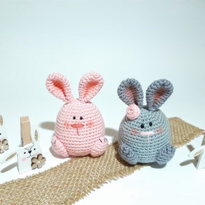 May include: Two crocheted bunny plushes, one pink and one gray, sitting on a burlap surface. Both bunnies have embroidered eyes and noses.