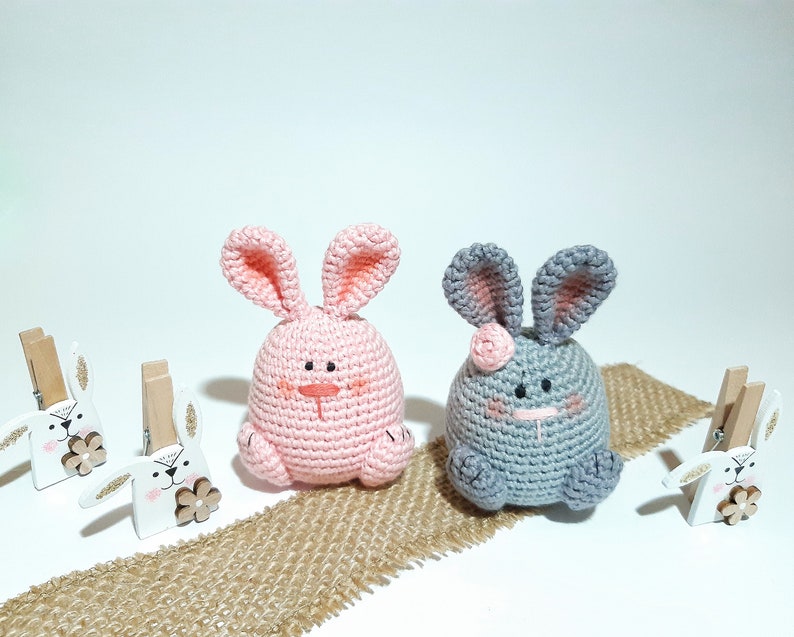 May include: Two crocheted bunny plushes, one pink and one gray, sitting on a burlap surface. Both bunnies have embroidered eyes and noses.