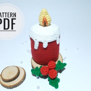 Op de afbeelding: Een gehaakte kaars met een rode basis, witte wasdruppels en een gele vlam. De kaars staat op een houten schijf met een takje groene bladeren en rode bessen. De tekst "PATTERN PDF" is zichtbaar.