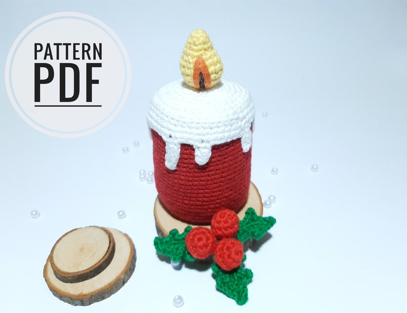 Op de afbeelding: Een gehaakte kaars met een rode basis, witte wasdruppels en een gele vlam. De kaars staat op een houten schijf met een takje groene bladeren en rode bessen. De tekst "PATTERN PDF" is zichtbaar.