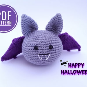 Puede incluir: Patrón de amigurumi de murciélago de crochet. El murciélago es gris con alas moradas y una cara linda. El patrón está disponible para descargar en PDF. "Feliz Halloween" está escrito en texto morado debajo del murciélago.