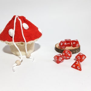 Pode incluir: Um saco de dados de croch&ecirc; em forma de cogumelo vermelho e branco com contas de madeira brancas nos cord&otilde;es. O saco est&aacute; cheio de dados vermelhos com n&uacute;meros brancos. Os dados est&atilde;o sobre uma base de madeira.