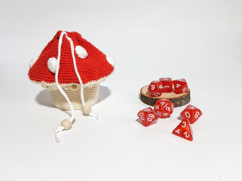 Pode incluir: Um saco de dados de croch&ecirc; em forma de cogumelo vermelho e branco com contas de madeira brancas nos cord&otilde;es. O saco est&aacute; cheio de dados vermelhos com n&uacute;meros brancos. Os dados est&atilde;o sobre uma base de madeira.
