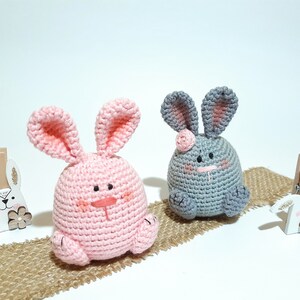 May include: Two crocheted bunny plushes, one pink and one gray, sitting on a burlap surface. Both bunnies have embroidered eyes and noses.