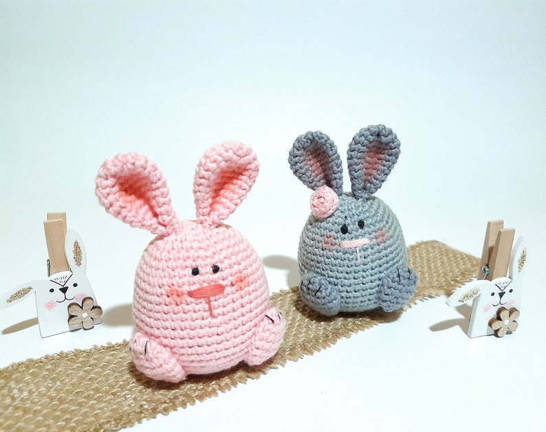 May include: Two crocheted bunny plushes, one pink and one gray, sitting on a burlap surface. Both bunnies have embroidered eyes and noses.