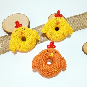 May include: Three crocheted chick toys in the shape of donuts. Two are yellow with red beaks and the other is orange with a red beak.