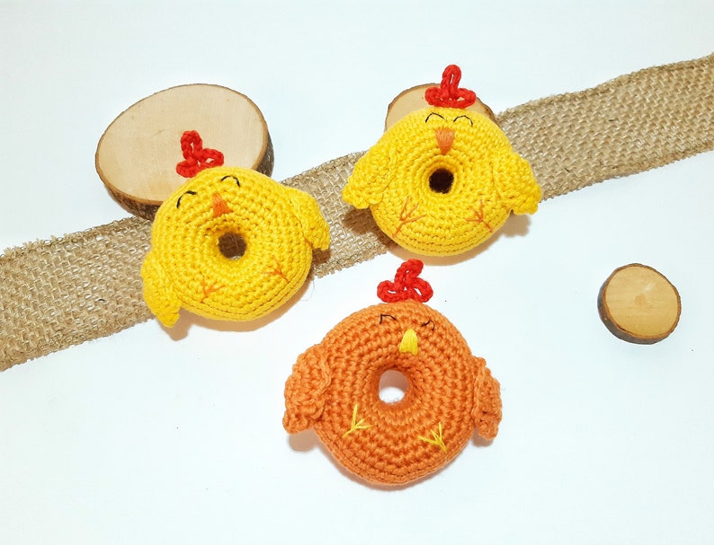 May include: Three crocheted chick toys in the shape of donuts. Two are yellow with red beaks and the other is orange with a red beak.