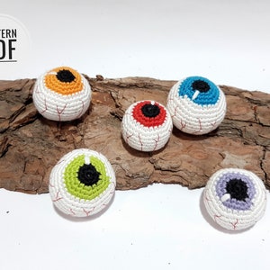 May include: Five crocheted eyeball ornaments with different colored pupils. The eyeballs are white with red veins and are sitting on a piece of tree bark.
