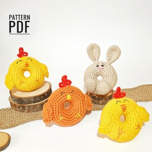 May include: Three crocheted amigurumi toys: a yellow chick, an orange chick, and a beige bunny. Each toy is shaped like a donut with a hole in the center. The toys are sitting on a wooden surface with a burlap ribbon.