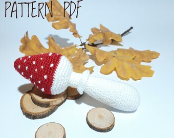 Può includere: Giocattolo a forma di fungo all'uncinetto con un cappello rosso e bianco e un gambo bianco. Il giocattolo è appoggiato su una superficie di legno con una foglia sullo sfondo. Il testo "PATTERN PDF" è visibile nell'angolo in alto a sinistra dell'immagine.