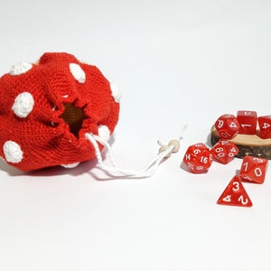 Pode incluir: Uma bolsa de dados de croch&ecirc; vermelha com bolinhas brancas e um cord&atilde;o de ajuste. A bolsa est&aacute; aberta e os dados vermelhos com n&uacute;meros brancos est&atilde;o caindo.