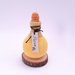 Puede incluir: Una botella de poci&oacute;n de crochet amarilla y naranja con una cuerda negra y una etiqueta que dice "Poci&oacute;n". La botella est&aacute; sentada sobre una rodaja de madera.