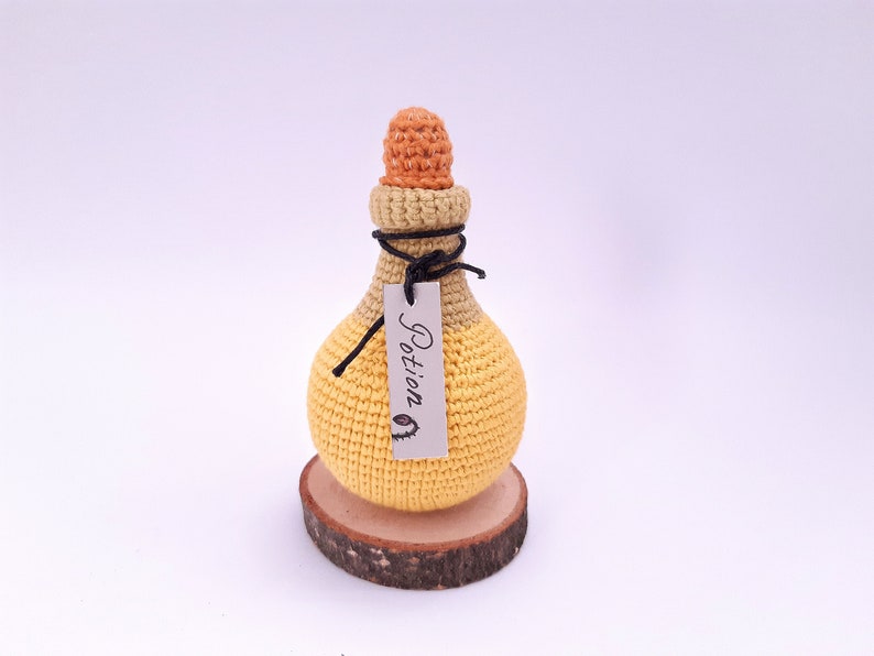 Puede incluir: Una botella de poci&oacute;n de crochet amarilla y naranja con una cuerda negra y una etiqueta que dice "Poci&oacute;n". La botella est&aacute; sentada sobre una rodaja de madera.
