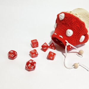 Pode incluir: Um saco de dados de croch&ecirc; vermelho e branco em forma de cogumelo com bolinhas brancas. Seis dados vermelhos com n&uacute;meros brancos est&atilde;o espalhados ao redor do saco.