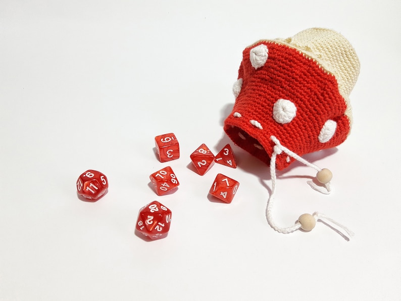 Pode incluir: Um saco de dados de croch&ecirc; vermelho e branco em forma de cogumelo com bolinhas brancas. Seis dados vermelhos com n&uacute;meros brancos est&atilde;o espalhados ao redor do saco.