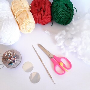 Op de afbeelding: Een verzameling knutselbenodigdheden, waaronder witte, gele, rode en groene bollen garen, witte vulling, een speldenkussen met spelden, twee zilveren cirkels, een haaknaald en een roze schaar. Voor creatieve projecten.