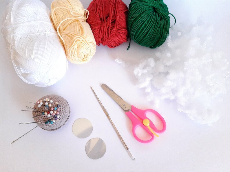 Op de afbeelding: Een verzameling knutselbenodigdheden, waaronder witte, gele, rode en groene bollen garen, witte vulling, een speldenkussen met spelden, twee zilveren cirkels, een haaknaald en een roze schaar. Voor creatieve projecten.