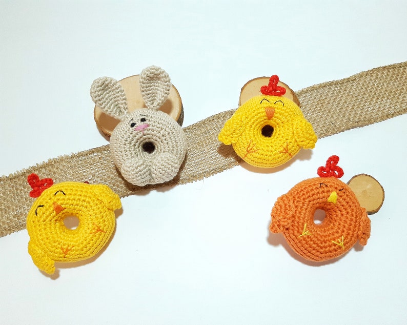 May include: Three crocheted stuffed animals: a bunny, a yellow chick, and an orange chick. The bunny is beige and has a pink nose. The chicks have red beaks and feet. All three animals are shaped like donuts.