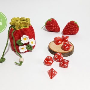 May include: A red crocheted drawstring bag with white flowers and green leaves. The bag is filled with red dice and is sitting on a wooden surface. There are two red crocheted strawberries next to the bag.