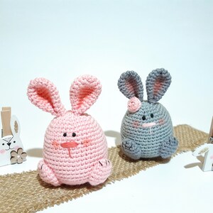 May include: Two crocheted bunny plushies, one pink and one gray, sitting on a burlap surface. Both bunnies have embroidered faces and pink ears.