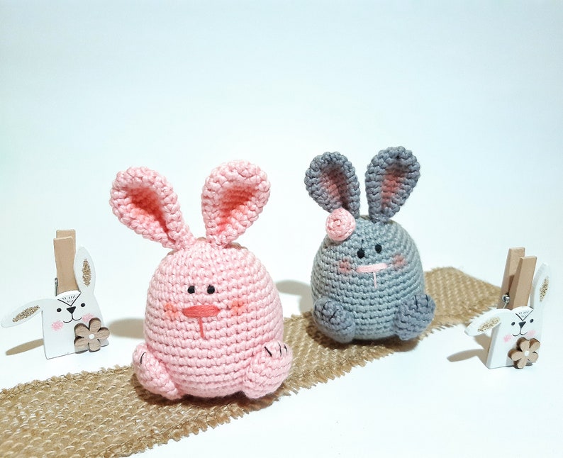 May include: Two crocheted bunny plushies, one pink and one gray, sitting on a burlap surface. Both bunnies have embroidered faces and pink ears.