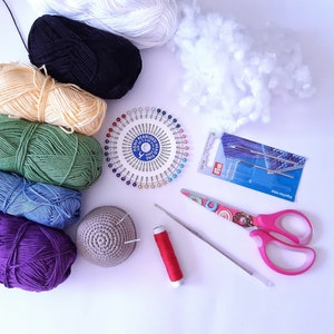 K&ouml;nnte beinhalten: Eine Sammlung bunter Wollkn&auml;uel, ein Nadelkissen mit Nadeln, eine kleine geh&auml;kelte M&uuml;tze, eine Schere, eine Spule roten Fadens und eine Packung Nadeln.
