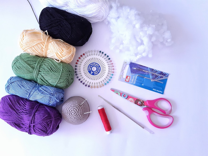 K&ouml;nnte beinhalten: Eine Sammlung bunter Wollkn&auml;uel, ein Nadelkissen mit Nadeln, eine kleine geh&auml;kelte M&uuml;tze, eine Schere, eine Spule roten Fadens und eine Packung Nadeln.