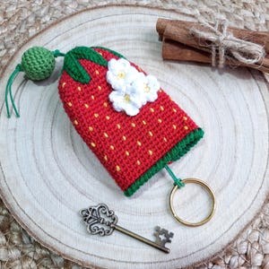 May include: A crocheted strawberry keyring with white flowers and a green stem. The keyring has a gold key ring and a small green ball charm.