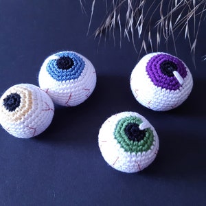 K&ouml;nnte beinhalten: Vier geh&auml;kelte Augapfel-Ornamente in verschiedenen Farben. Die Aug&auml;pfel sind wei&szlig; mit roten N&auml;hten und haben unterschiedlich farbige Iris: blau, lila, gr&uuml;n und gelb.