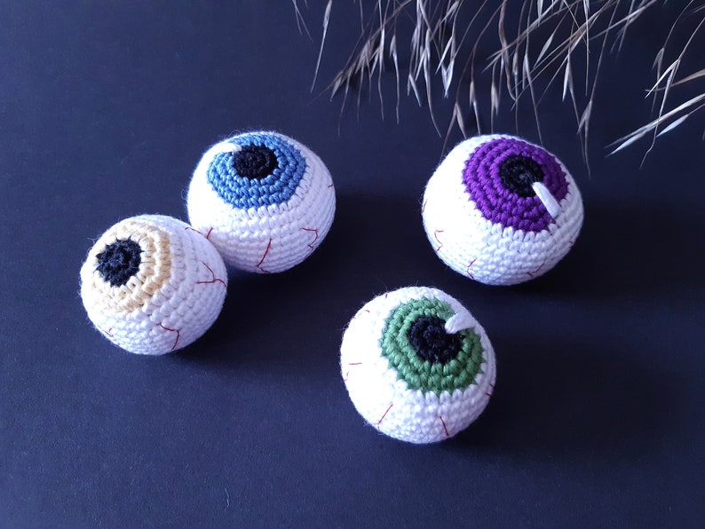 K&ouml;nnte beinhalten: Vier geh&auml;kelte Augapfel-Ornamente in verschiedenen Farben. Die Aug&auml;pfel sind wei&szlig; mit roten N&auml;hten und haben unterschiedlich farbige Iris: blau, lila, gr&uuml;n und gelb.