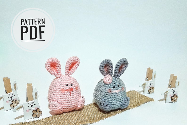 May include: Two crocheted bunny plushies, one pink and one gray, sitting on a burlap surface. The bunnies have embroidered faces and are holding a pink flower.