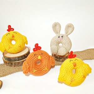 May include: Four crocheted stuffed animals in the shape of chicks and a bunny. The chicks are yellow, orange, and yellow with a red beak and feet. The bunny is beige with a pink nose and a red nose. The animals are sitting on a burlap surface.