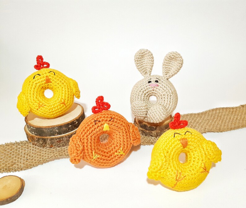 May include: Four crocheted stuffed animals in the shape of chicks and a bunny. The chicks are yellow, orange, and yellow with a red beak and feet. The bunny is beige with a pink nose and a red nose. The animals are sitting on a burlap surface.