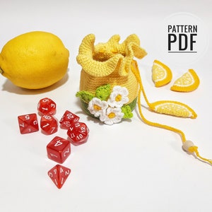 May include: A crocheted lemon-shaped dice bag with a drawstring closure and a daisy design. The bag is yellow and white and has a wooden bead on the drawstring. There are red dice scattered around the bag.