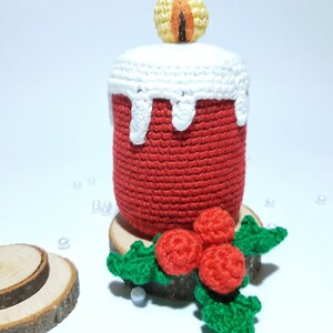 Op de afbeelding: Een handgemaakte gehaakte kaars in rood met witte wasdruppeldetails. De kaars heeft een gele en oranje vlam en staat op een houten voet met een hulsttak. Een feestelijk, decoratief item.