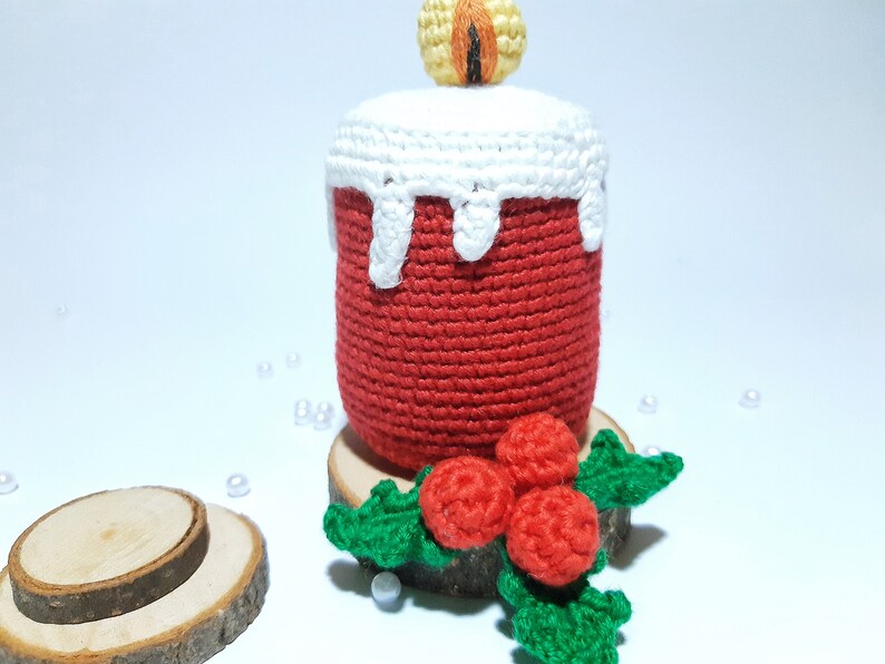 Op de afbeelding: Een handgemaakte gehaakte kaars in rood met witte wasdruppeldetails. De kaars heeft een gele en oranje vlam en staat op een houten voet met een hulsttak. Een feestelijk, decoratief item.