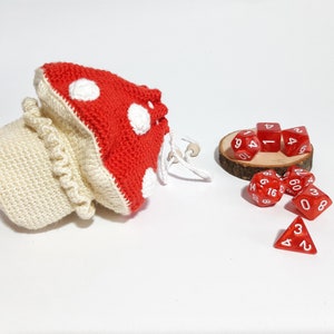 Pode incluir: Um saco de dados de croch&ecirc; em forma de cogumelo com detalhes em branco e vermelho. O saco est&aacute; cheio de dados vermelhos com n&uacute;meros brancos.
