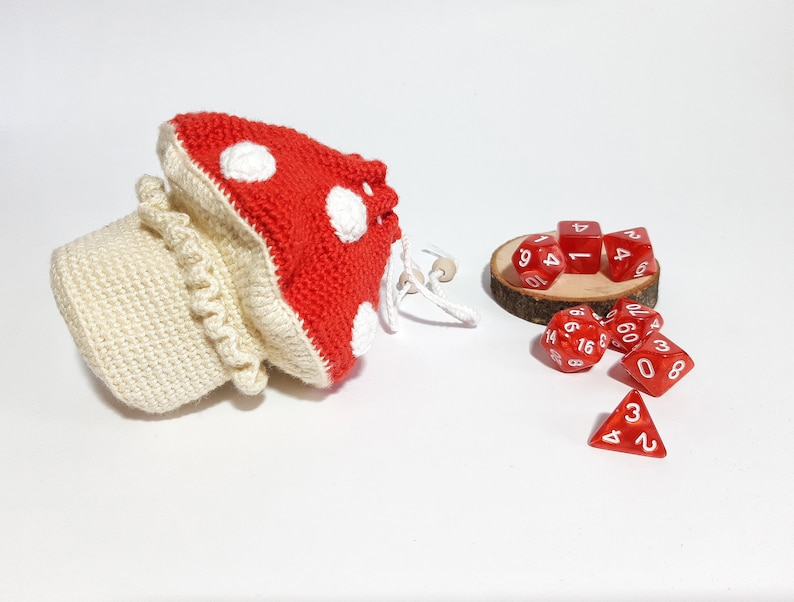 Pode incluir: Um saco de dados de croch&ecirc; em forma de cogumelo com detalhes em branco e vermelho. O saco est&aacute; cheio de dados vermelhos com n&uacute;meros brancos.