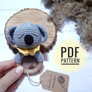 Op de afbeelding: Gehaakte koala rammelaar met een houten ring. De koala is grijs met zwarte ogen en een gele kraag. Het speelgoed is handgemaakt en wordt geleverd met een PDF-patroon.