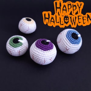 K&ouml;nnte beinhalten: Vier geh&auml;kelte Aug&auml;pfel-Ornamente in verschiedenen Farben, darunter Gr&uuml;n, Lila, Blau und Beige. Die Aug&auml;pfel befinden sich auf einem schwarzen Hintergrund mit dem Text "Happy Halloween" in Orange.