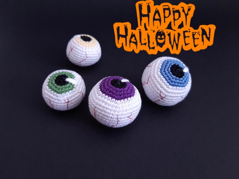K&ouml;nnte beinhalten: Vier geh&auml;kelte Aug&auml;pfel-Ornamente in verschiedenen Farben, darunter Gr&uuml;n, Lila, Blau und Beige. Die Aug&auml;pfel befinden sich auf einem schwarzen Hintergrund mit dem Text "Happy Halloween" in Orange.